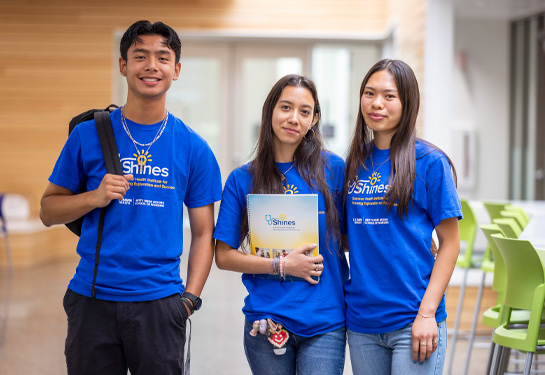 Three students stand together, one holding backpack, one holding folder, all smiling at camera