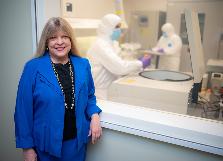 Professor Jan Nolta wearing a black shirt, blue jacket and a long necklace, is standing next to window. In the background, two researchers in white protective overalls and blue masks work in the GMP facility.