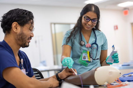 Nursing student in green scrubs practices on mannequin arm while nursing instructor in blue scrubs looks on